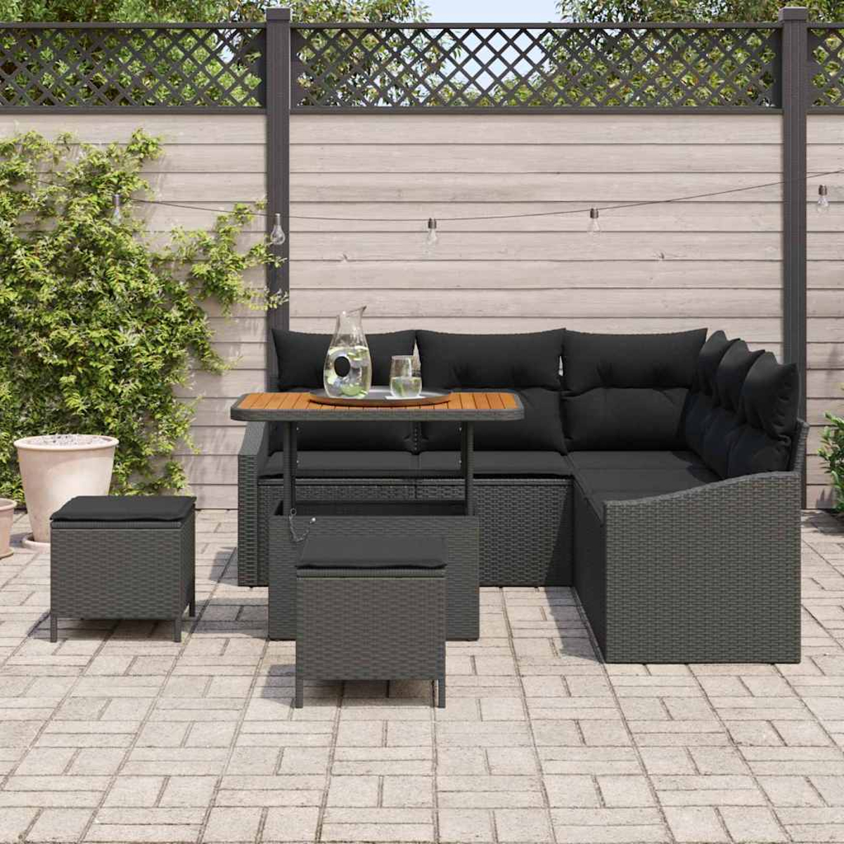 Garten-Terrassenset mit grauem Korbsessel, Tisch und Hockern. Schwarze Kissen. Im Hintergrund ein Zaun und Grün.