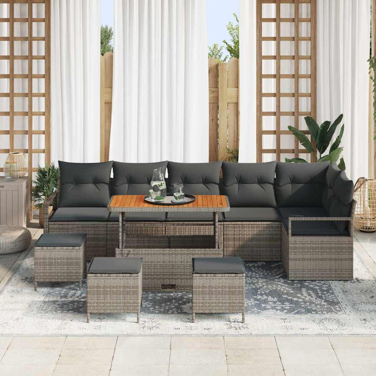 Sofá seccional para exteriores con cojines grises y mesa de madera. Incluye otomanas a juego sobre una alfombra estampada, cortinas y valla al fondo.
