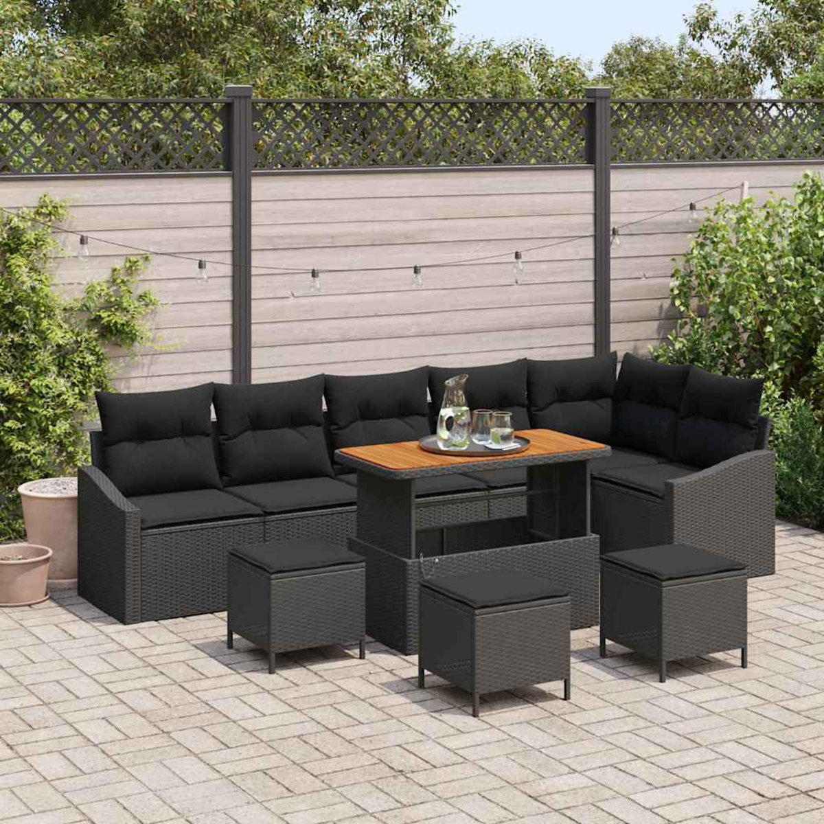 Terrassengarnitur mit schwarzen Kissen, Holztisch und grauen Korbmöbeln. Beinhaltet Sitzgelegenheiten, Hocker und Lichterketten.