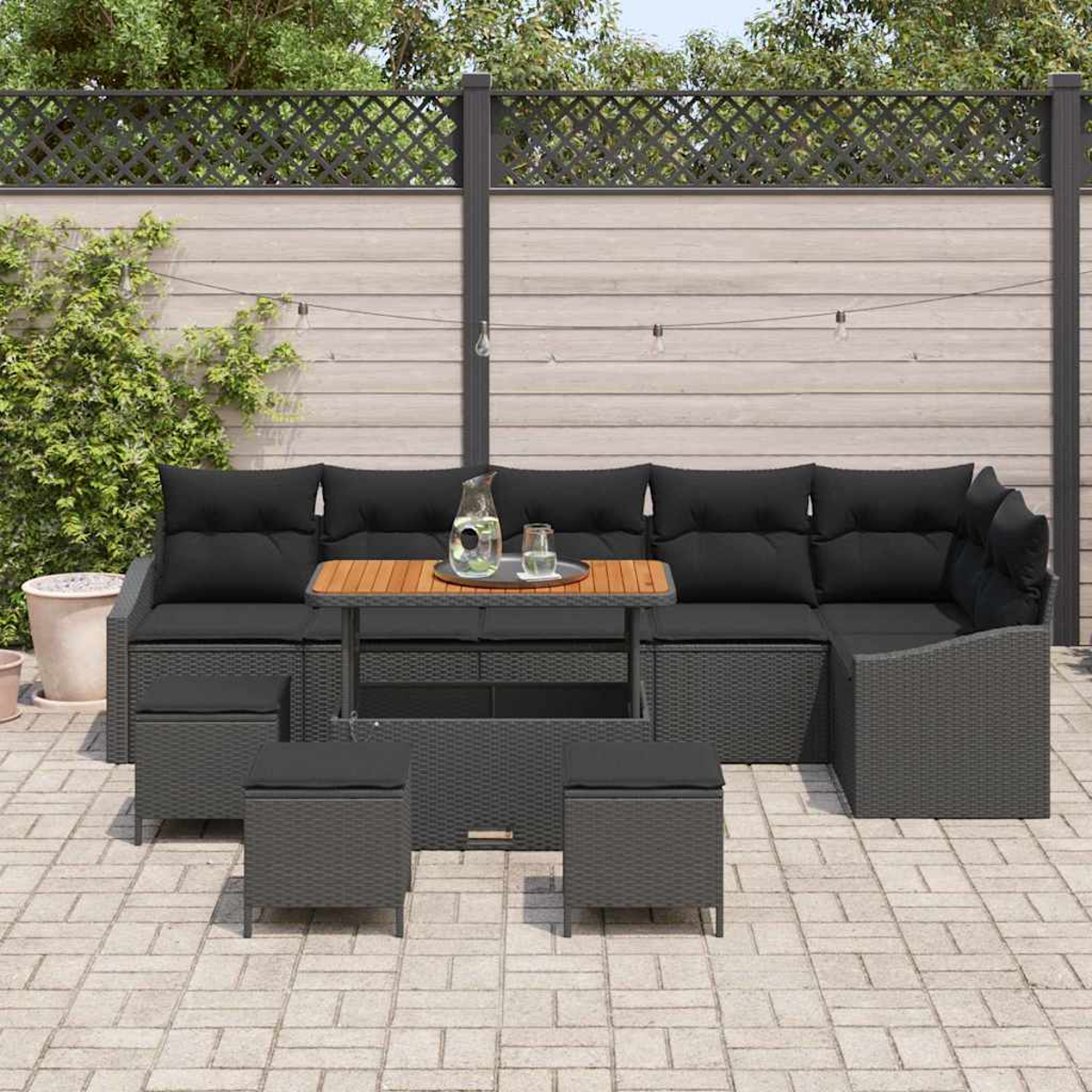 Outdoor-Ecksofa mit schwarzen Kissen, Holztisch und zwei Hockern. Eine Wasserkaraffe und ein Glas auf dem Tisch. Im Garten.