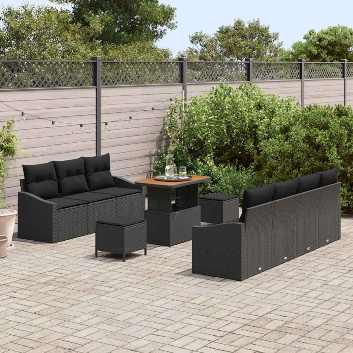 Terrassenmöbel. Schwarze Korbsessel, Hocker und Tisch auf Steinterrasse, Zaun und Grün im Hintergrund.