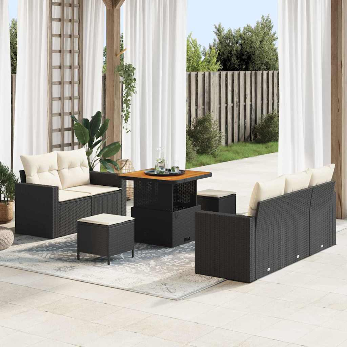 Set patio esterno con mobili in vimini neri, cuscini beige e un tavolo in legno, su un patio con tende bianche e una recinzione.