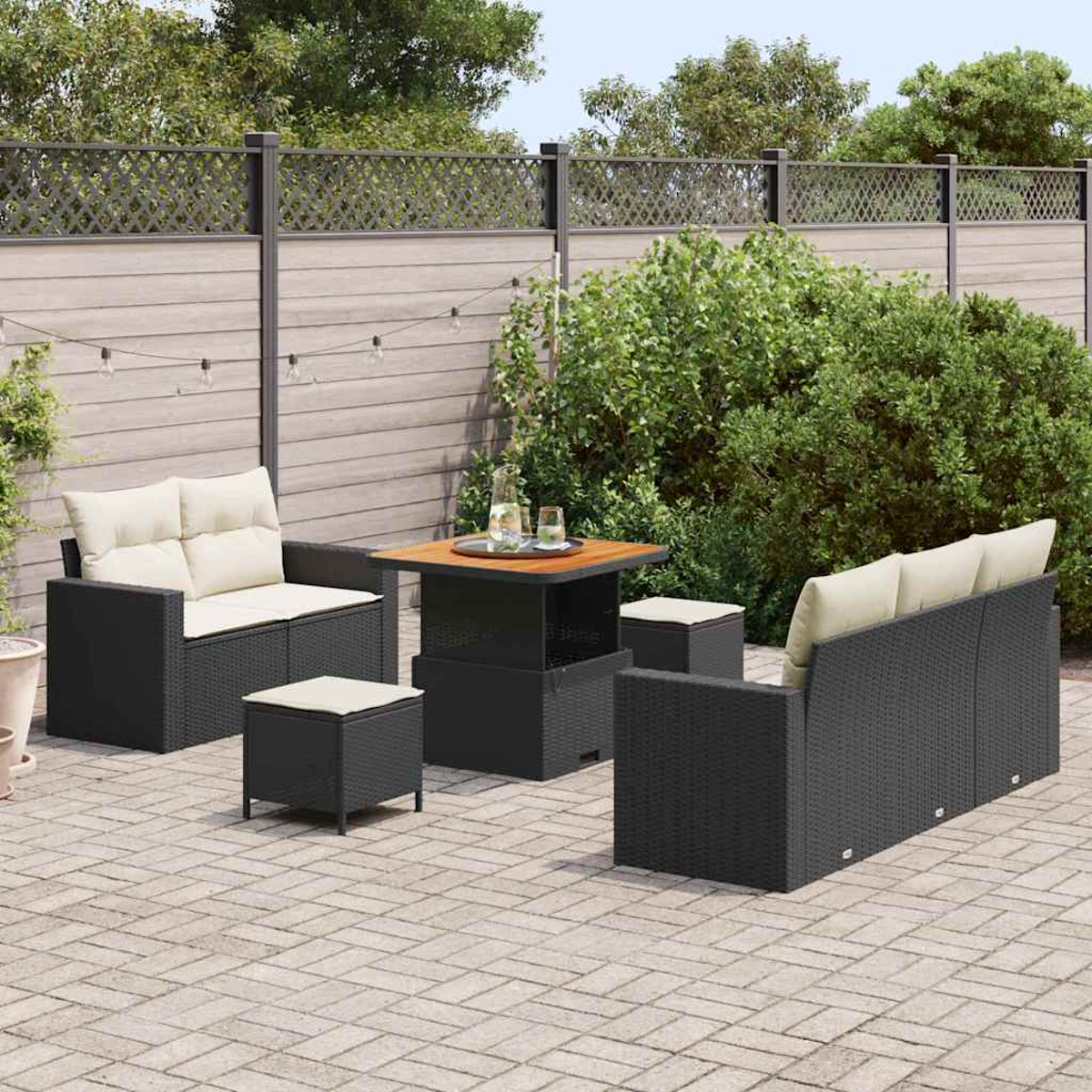 Set di mobili da patio. Sedie e divano neri intrecciati con cuscini beige e un tavolo con piano in legno. Piante verdi e recinzione sullo sfondo.