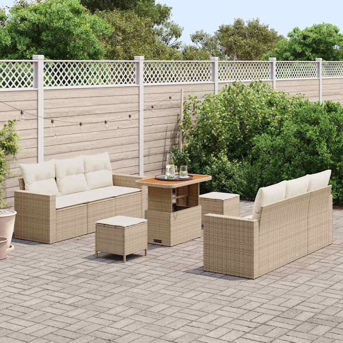 Terrassenmöbel-Set: Wicker-Sofa, Tisch und Ottomanen auf gepflasterter Terrasse. Holzzaun und Grün im Hintergrund.