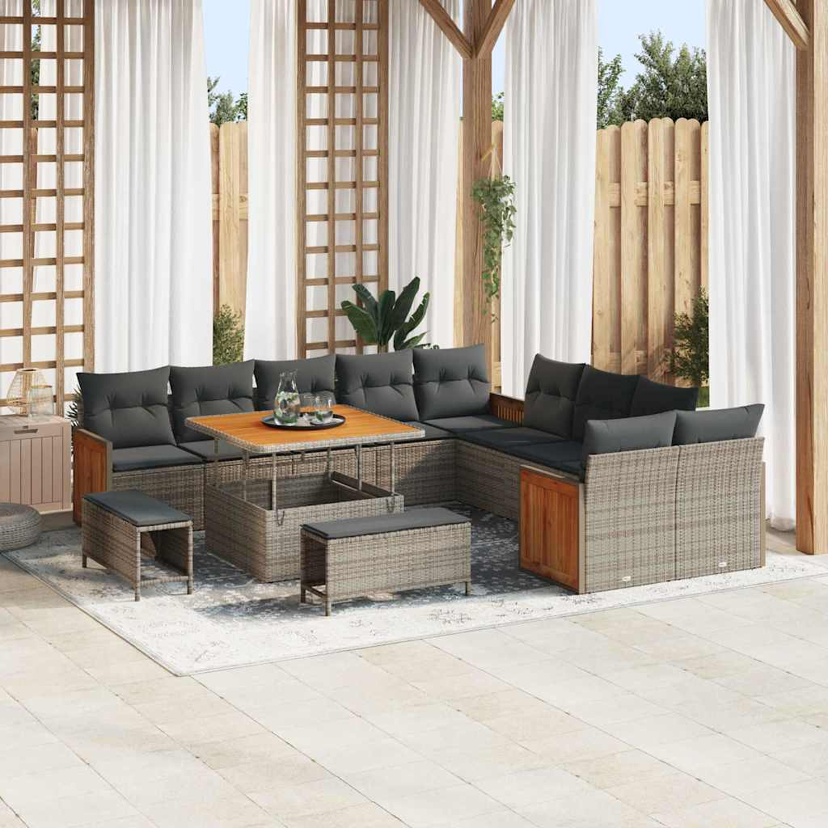 Set da patio esterno con cuscini grigi, tavolo in legno e panche. Tende bianche e recinzione in legno sullo sfondo.