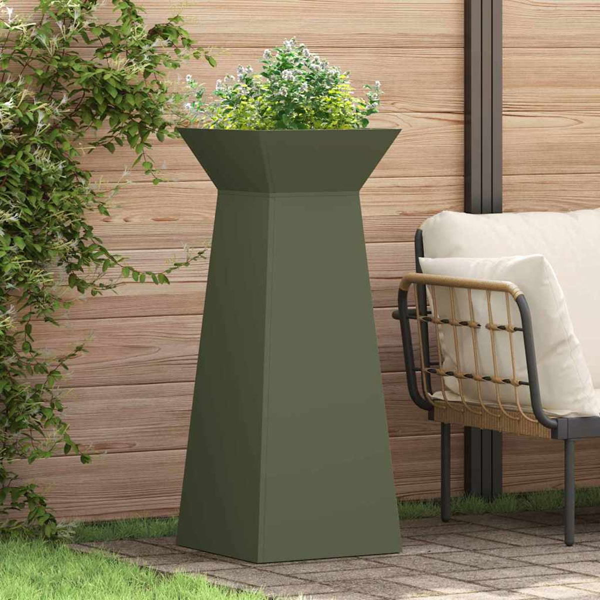 Un'alta fioriera verde oliva con fiori bianchi si trova su un patio. Una sedia con un cuscino color crema è accanto, con una recinzione di legno sullo sfondo.