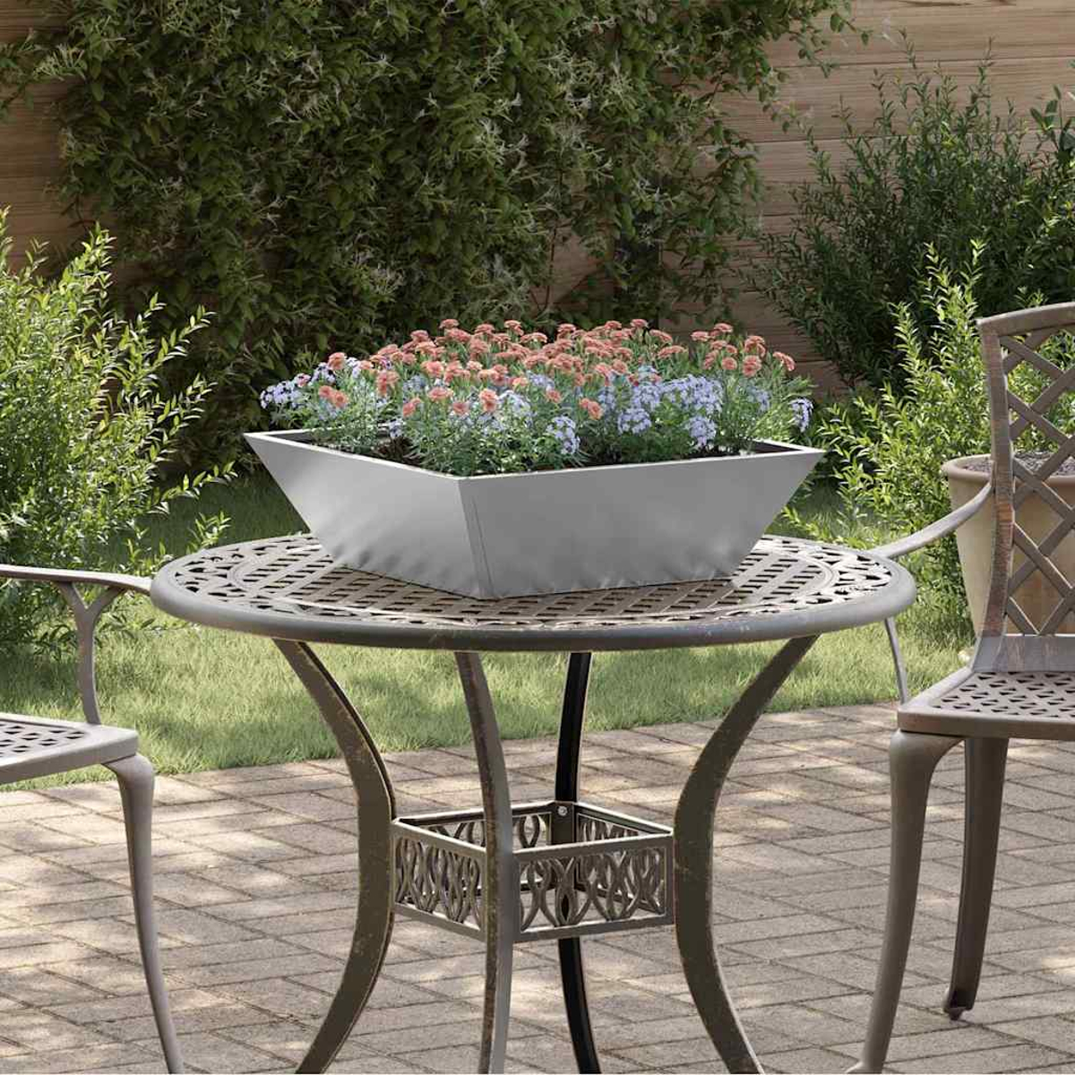 Un tavolo di metallo con una fioriera di fiori si trova su un patio. Arbusti verdi e una staccionata di legno sullo sfondo.