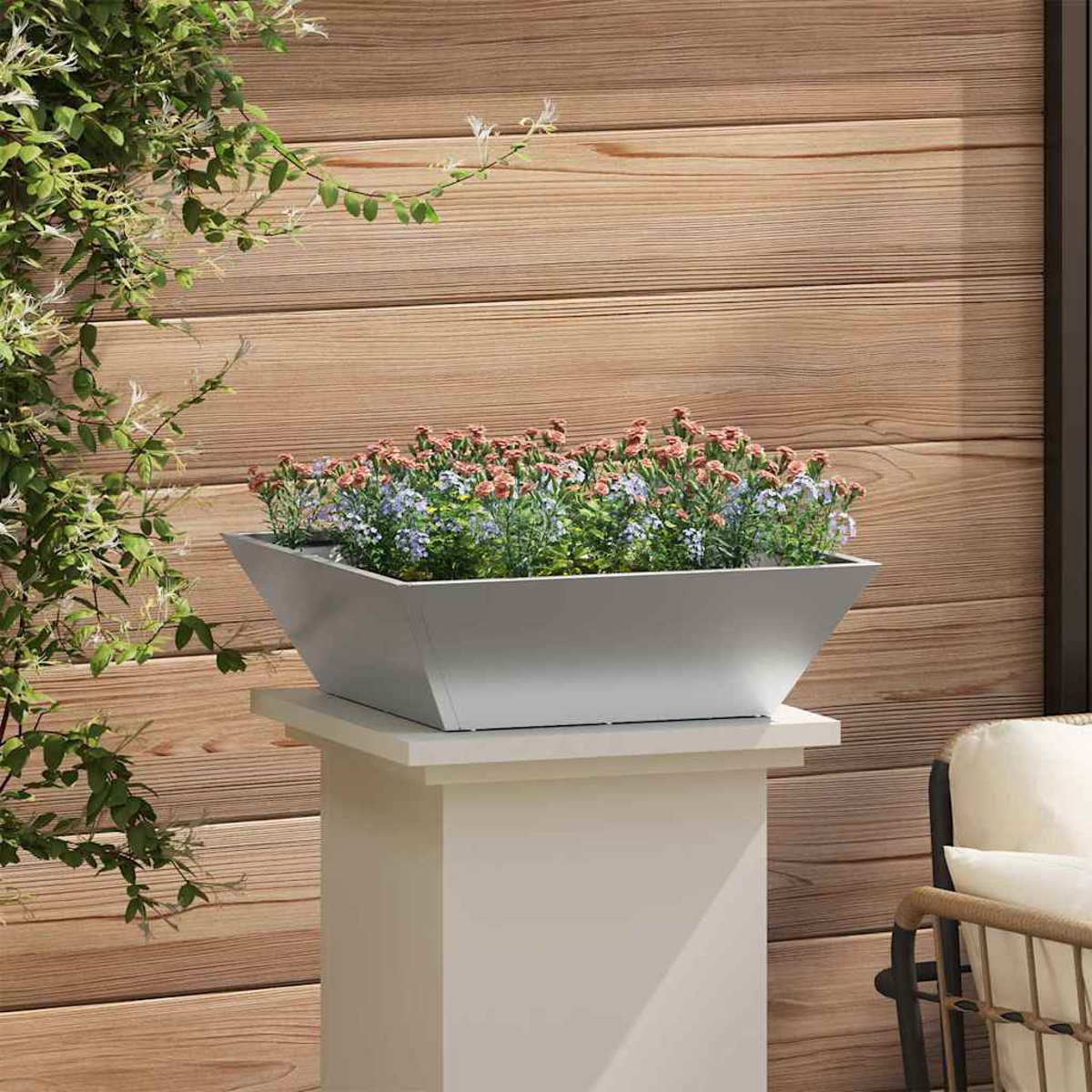 Una fioriera grigia piena di fiori è su un piedistallo bianco. Parete in legno sullo sfondo, con fogliame verde a sinistra.