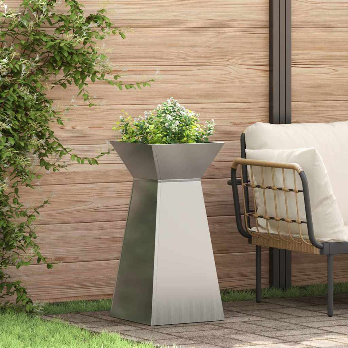 Una fioriera di metallo con piante si trova su un patio. Una sedia con un cuscino bianco è accanto a una recinzione di legno. È visibile erba verde.