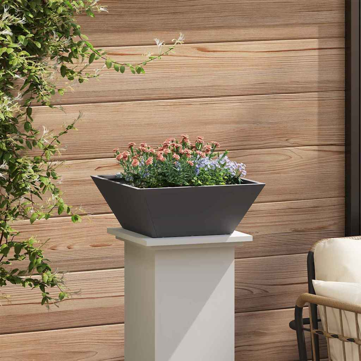 Una fioriera grigio scuro con fiori colorati poggia su un piedistallo bianco. Parete in legno sullo sfondo, con una sedia a destra.