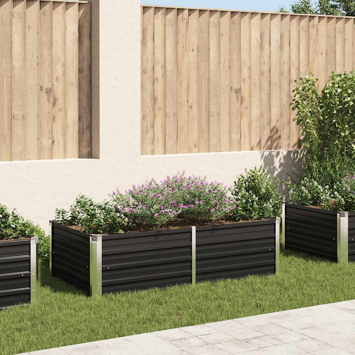 Drei schwarze Metallpflanzgefäße mit Blumen und Pflanzen auf grünem Gras, vor einem Holzzaun und einer weißen Wand.