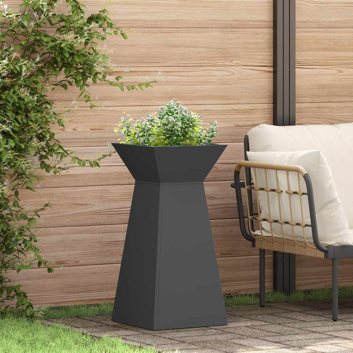 Una fioriera grigio scuro con piante è su un patio di pietra. C'è una sedia con un cuscino bianco dietro. Recinzione di legno sullo sfondo.