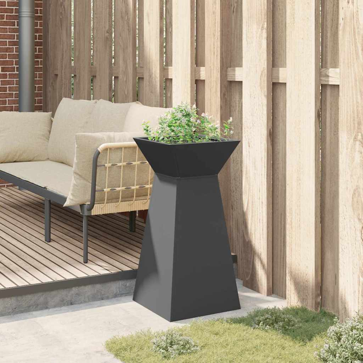 Un'alta fioriera grigio scuro con piante su un patio. Sfondo con recinzione in legno e divano beige. Erba in primo piano.