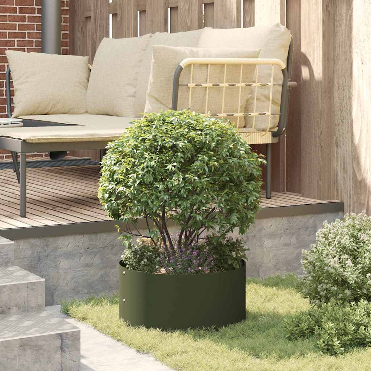 Un albero rotondo in una fioriera di metallo verde è sull'erba. Dietro l'albero c'è un set da patio con cuscini beige. Sono visibili mattoni e una recinzione di legno.