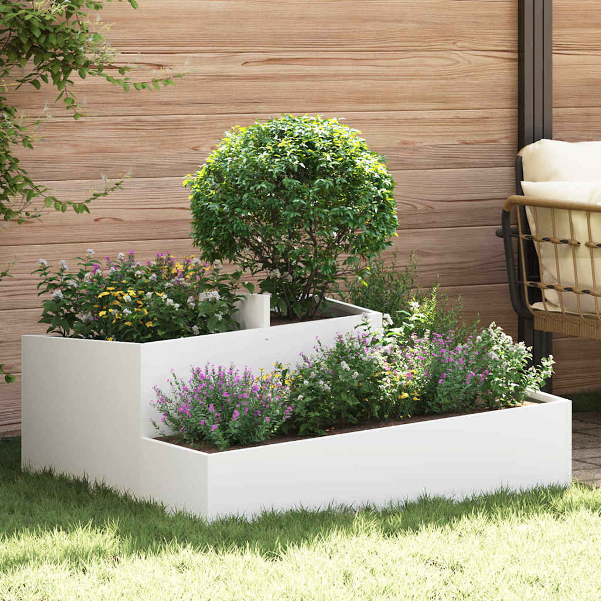 Una fioriera bianca a più livelli con fiori colorati, erba e un alberello. Una staccionata in legno e una sedia sullo sfondo.