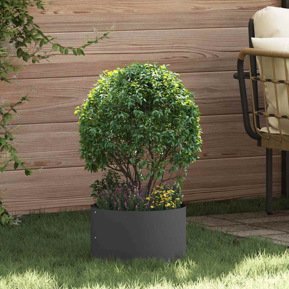 Una fioriera rotonda grigio scuro contiene un cespuglio verde con piccoli fiori viola e gialli alla base, in un giardino con pannelli di legno.