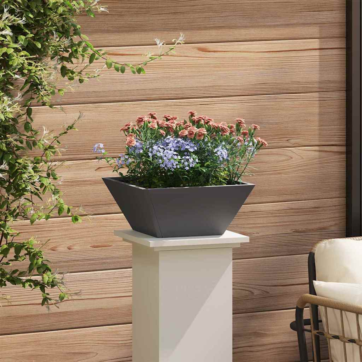 Una fioriera grigia con fiori rosa e blu poggia su un piedistallo bianco. Tavole di legno formano lo sfondo, con viti verdi a sinistra.