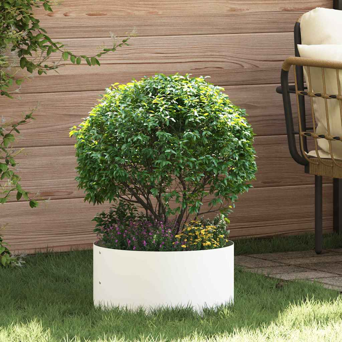 Una fioriera rotonda bianca contiene un albero verde e frondoso con fiori colorati. Lo sfondo mostra rivestimento in legno, erba e una sedia.