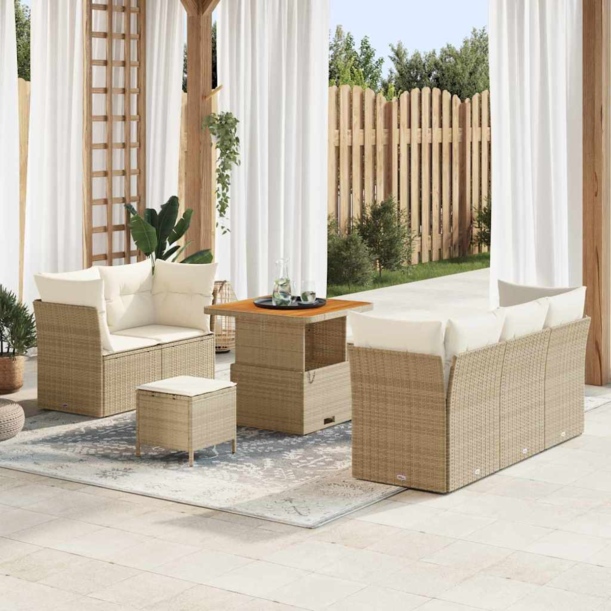 Terrassenset: Korbmöbel, Sofa, Stühle, Tisch und Hocker. Cremefarbene Kissen und Vorhänge. Holzzaun und Grün im Hintergrund.