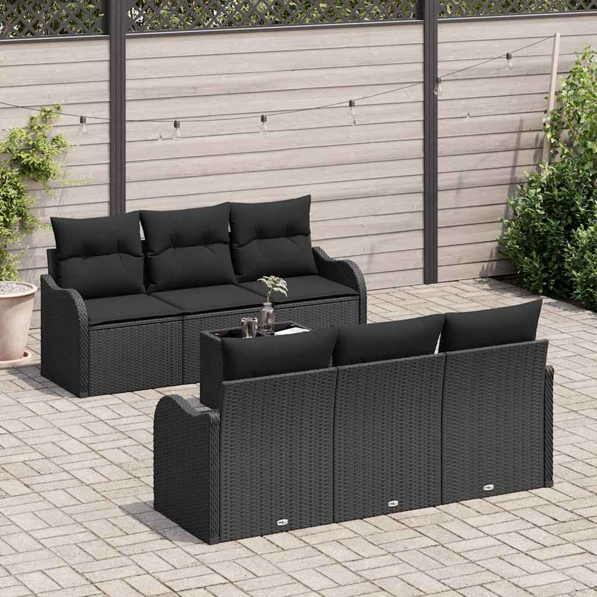 Szene auf der Außenterrasse mit schwarzem Korbmöbel-Set und Couchtisch. Holzzaun im Hintergrund, Lichterketten oben.