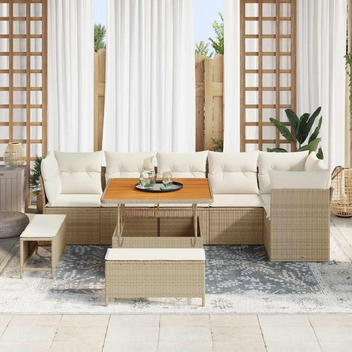 Set di sedute per esterni con cuscini beige e struttura in vimini. Include tavolo, panca e tappeto. Tende bianche e graticcio in legno sullo sfondo.