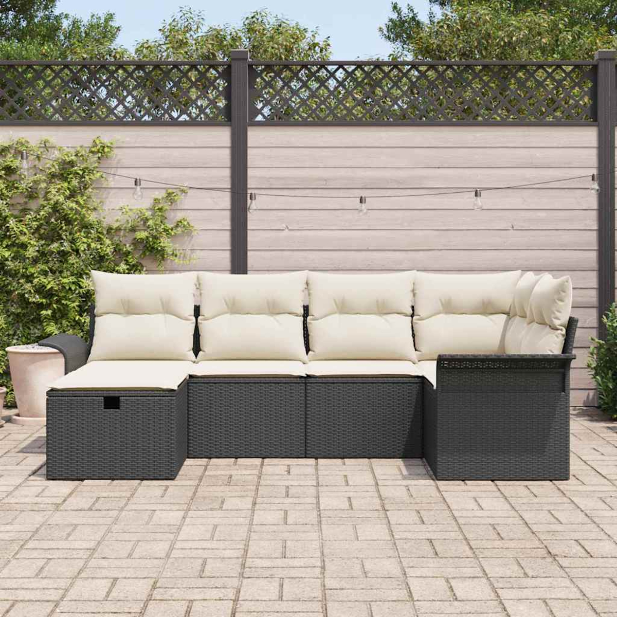 Schwarzes Outdoor-Rattan-Ecksofa mit cremefarbenen Kissen auf einer Terrasse. Im Hintergrund ein Zaun und Lichterketten.