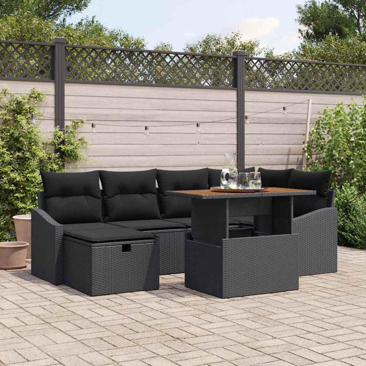 Schwarzes Outdoor-Korbmöbel-Set mit Tisch. Hinterhof mit Holzzaun und Lichterketten. Getränke auf dem Tisch. Gepflasterte Terrasse.
