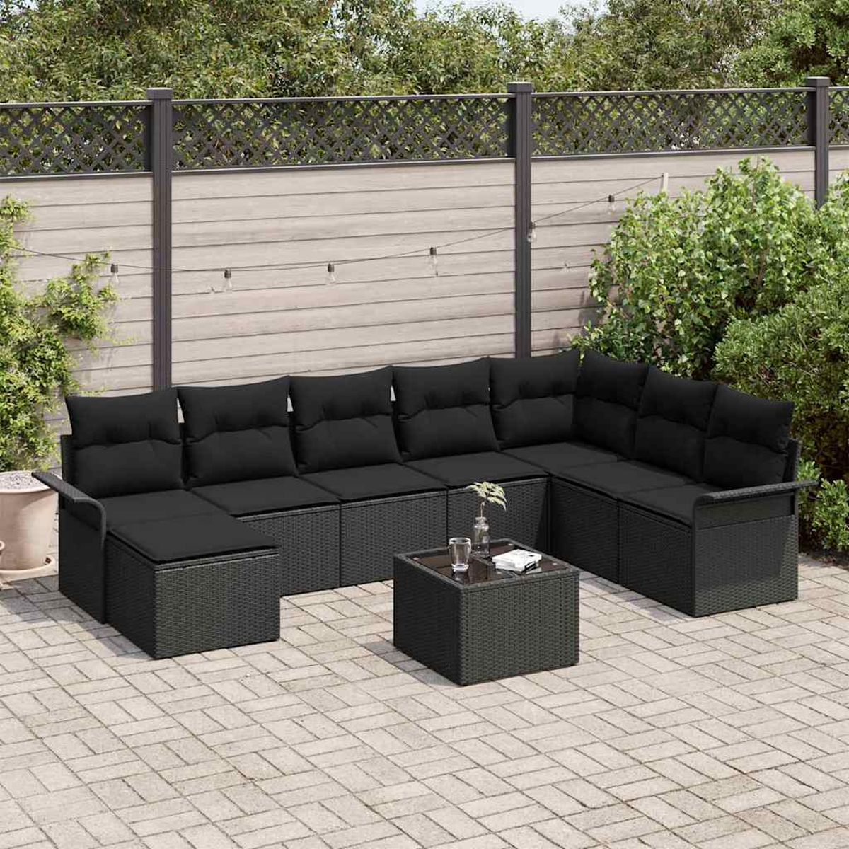 Garten-Terrassengarnitur. Schwarzes Ecksofa aus Korbgeflecht und quadratischer Tisch auf einer Ziegelstein-Terrasse. Im Hintergrund ein Zaun und Pflanzen.