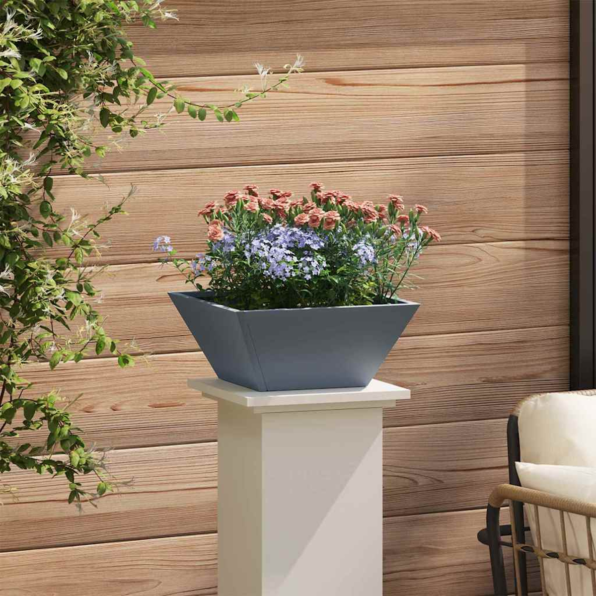 Una fioriera grigia su un piedistallo bianco contiene fiori rosa e viola. Sullo sfondo un muro di legno, con piante verdi a sinistra.