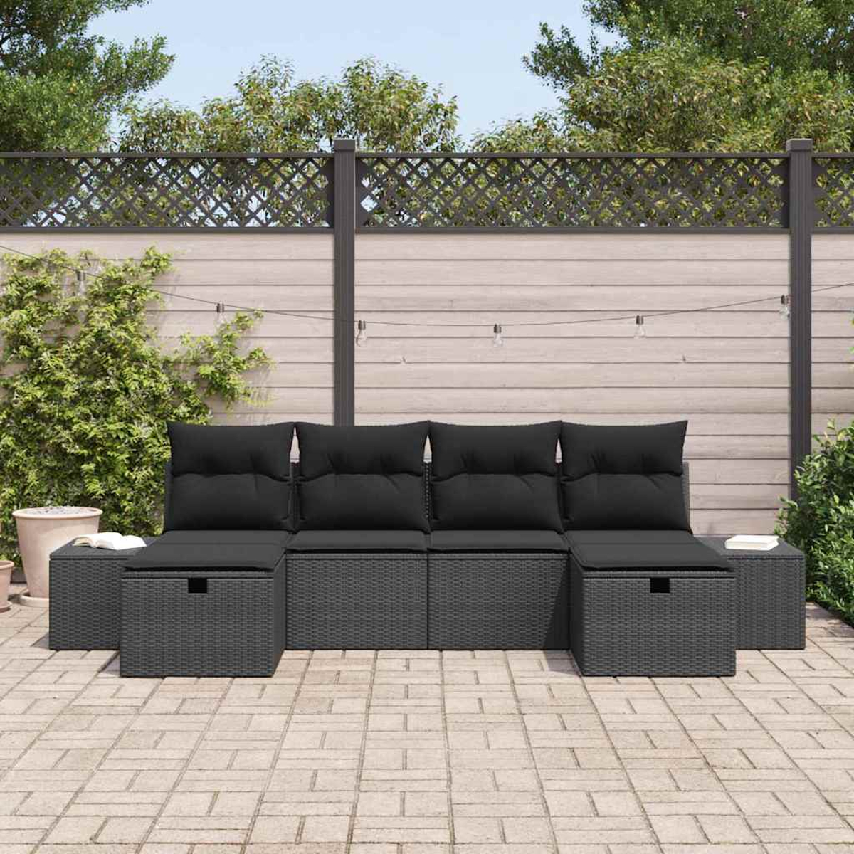 Graues Outdoor-Wicker-Ecksofa mit schwarzen Kissen auf einer Ziegelterrasse. Im Hintergrund ein Holzzaun und Grün.