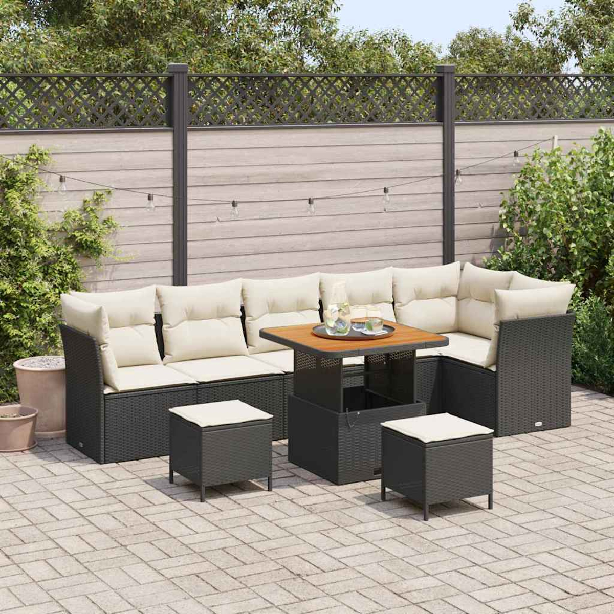Terrassen-Set: graues Korbmöbel mit cremefarbenen Kissen, quadratischer Tisch, zwei Hocker. Holzzaun, Lichterkette und Grün im Hintergrund.
