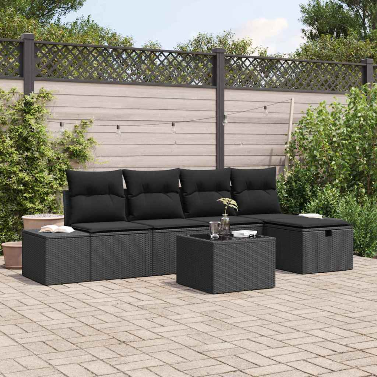 Outdoor-Sofa-Set aus schwarzem Korbgeflecht mit Kissen, Couchtisch und Ottomane auf einer gepflasterten Terrasse, Hintergrund ist ein Holzzaun und Grün.