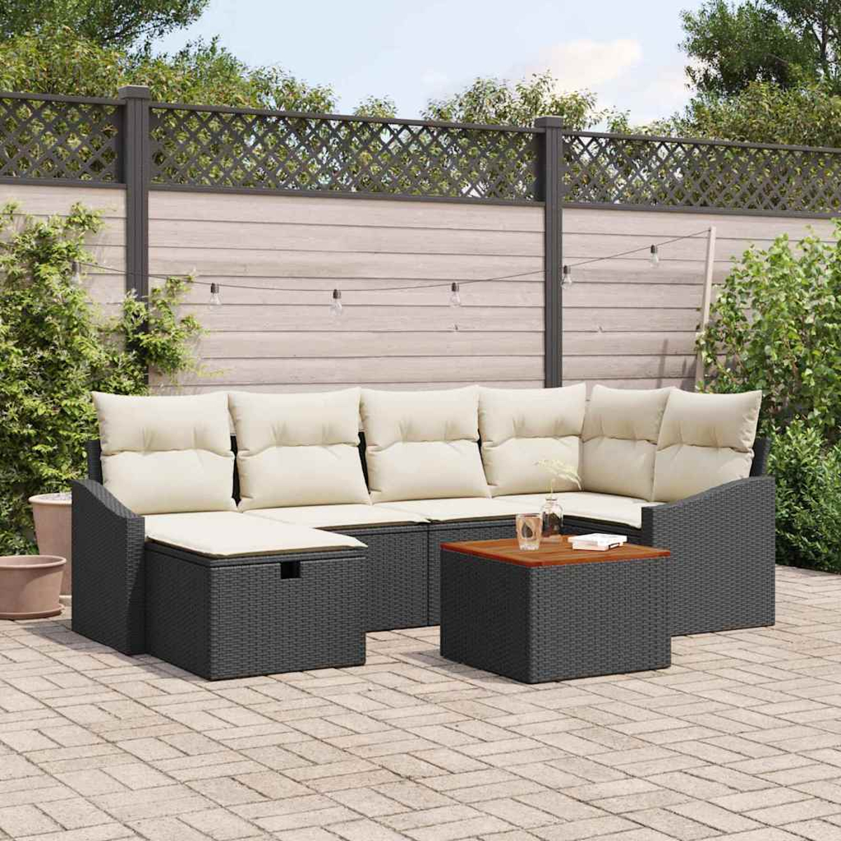 Outdoor-Ecksofa mit cremefarbenen Kissen, dunkelgrauem Korbgeflecht und einem kleinen Tisch. Der Hintergrund zeigt einen Zaun und Grün.