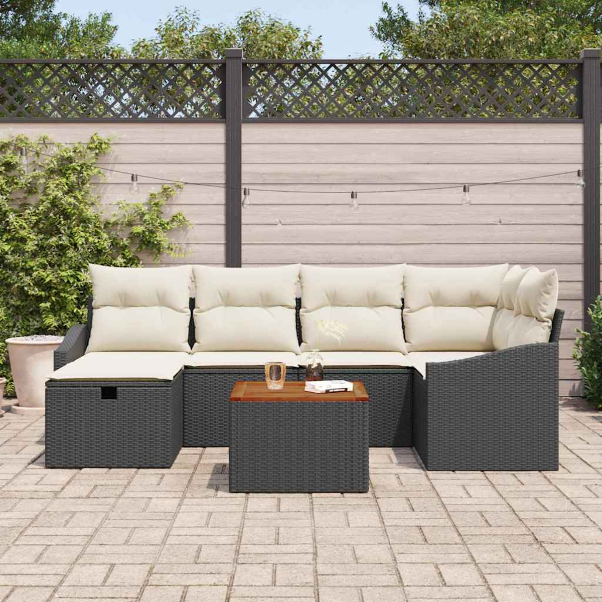 Garten-Sitzgruppe: graues Korbmöbel-Sofa mit cremefarbenen Kissen und einem Couchtisch auf einer Backstein-Terrasse, Holzzaun im Hintergrund.