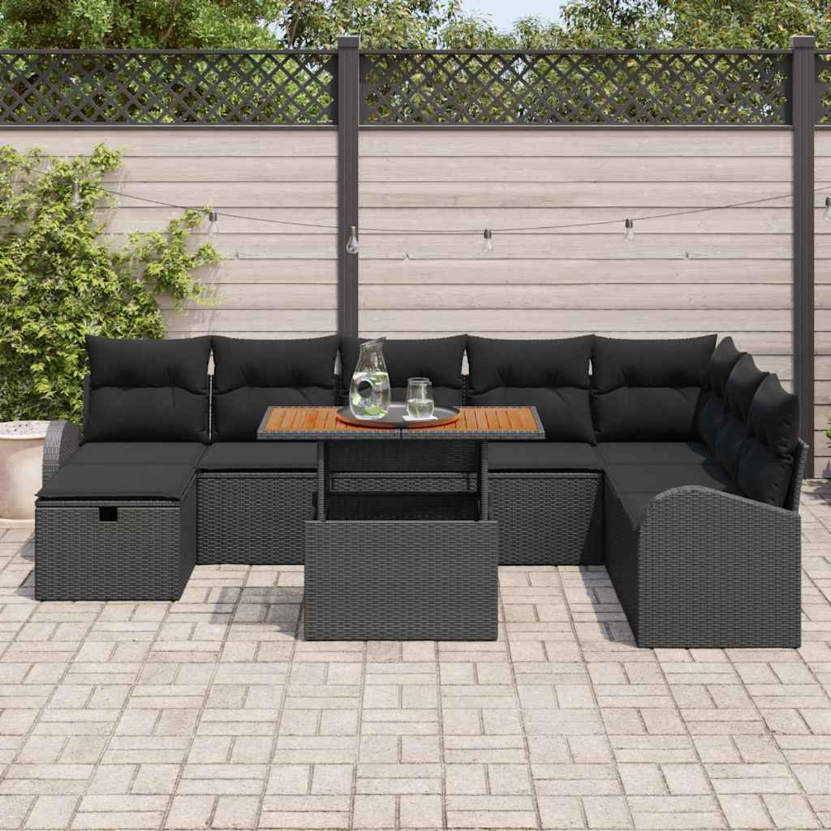 Ein Outdoor-Terrassenset mit grauen Korbmöbeln, schwarzen Kissen und einem Holztisch. Der Hintergrund zeigt einen Holzzaun und Grün.