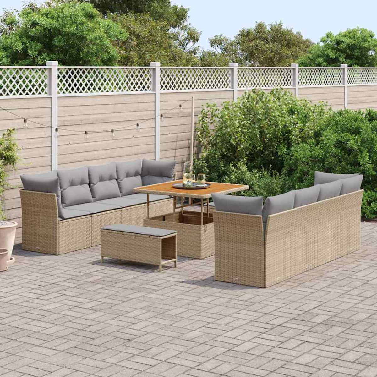 Außenbereich mit Tisch und Sofas. Beige Möbel auf einer Steinterrasse, mit weißem Zaun und grünen Bäumen im Hintergrund.