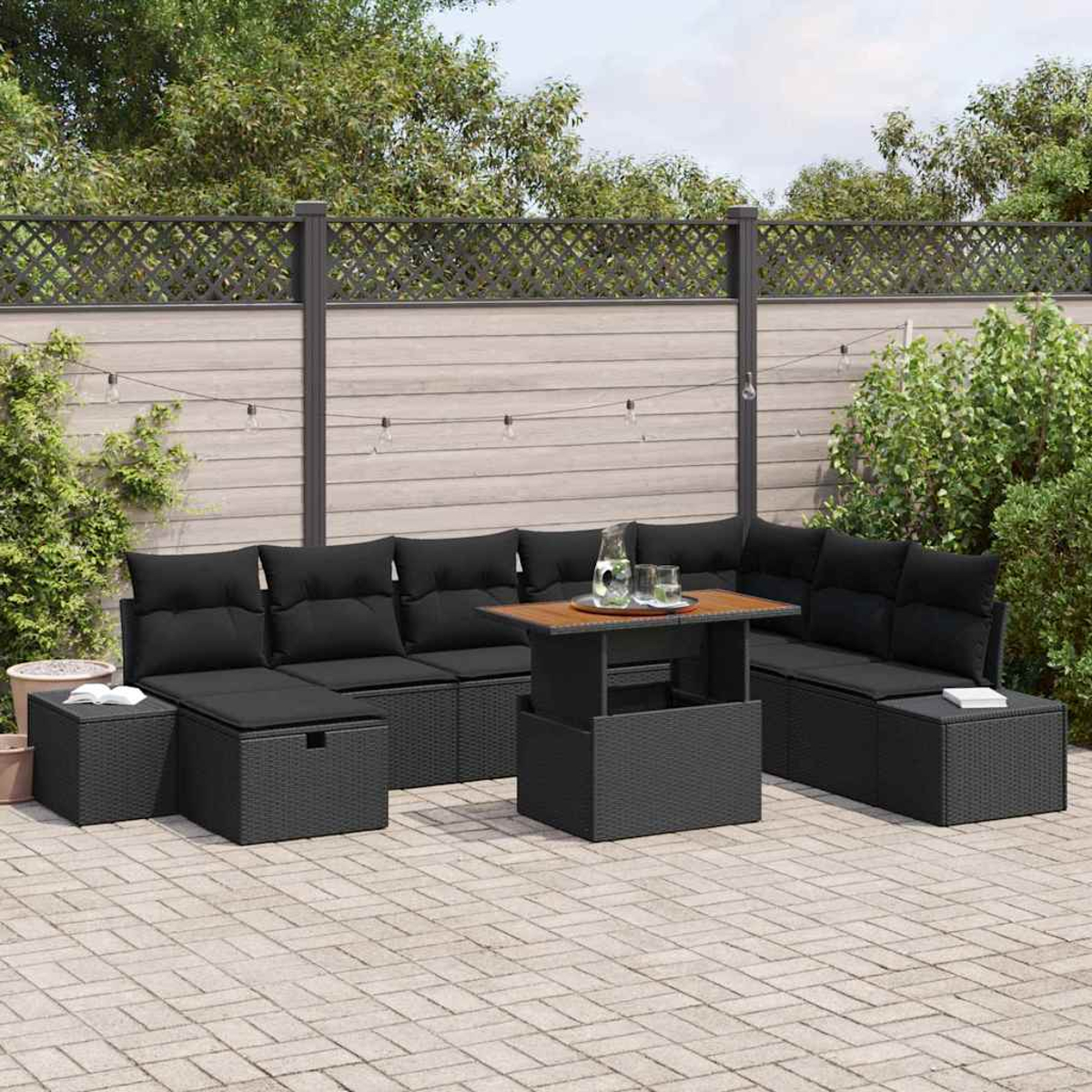 Schwarzes Outdoor-Korbmöbel-Set mit schwarzen Kissen, einem Holztisch und Grün im Hintergrund. Lichterketten hängen oben.