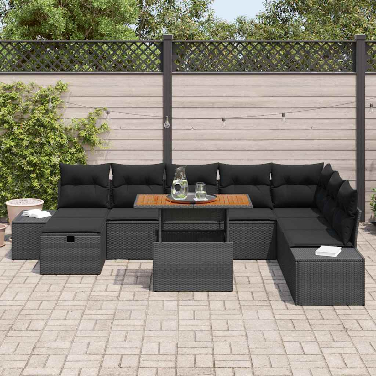 Graues Outdoor-Sofa aus Korbgeflecht mit schwarzen Kissen und einem Holztisch auf einer Steinterrasse. Im Hintergrund ein Zaun und Grün.