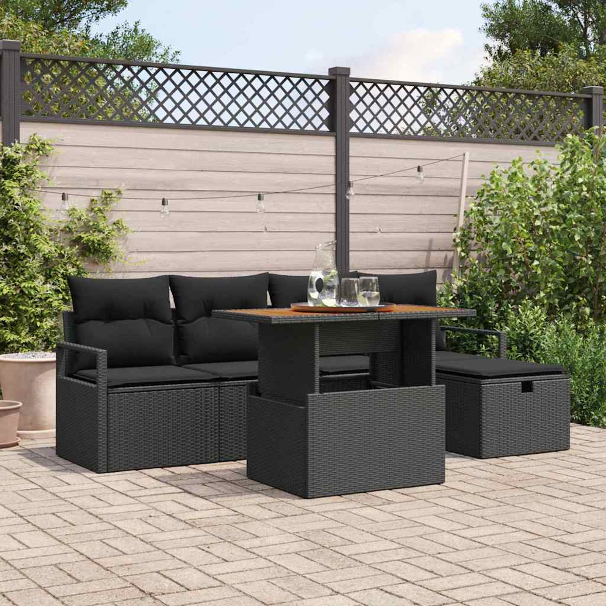 Garten-Terrassenset. Schwarze Korbmöbel, ein Tisch mit Glaskrug und Gläsern auf einer Steinterrasse. Ein Holzzaun ist im Hintergrund.