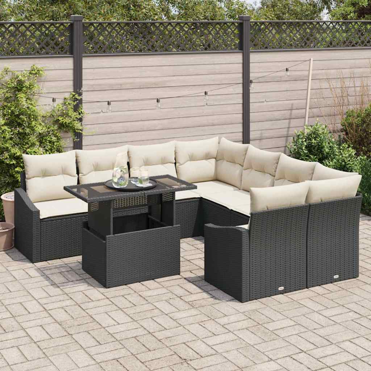 Garten-Sitzgruppe mit dunkelgrauem Korbgeflecht, beigen Kissen und einem Tisch mit Hebefunktion auf einer Terrasse. Hintergrund: Zaun und Pflanzen.