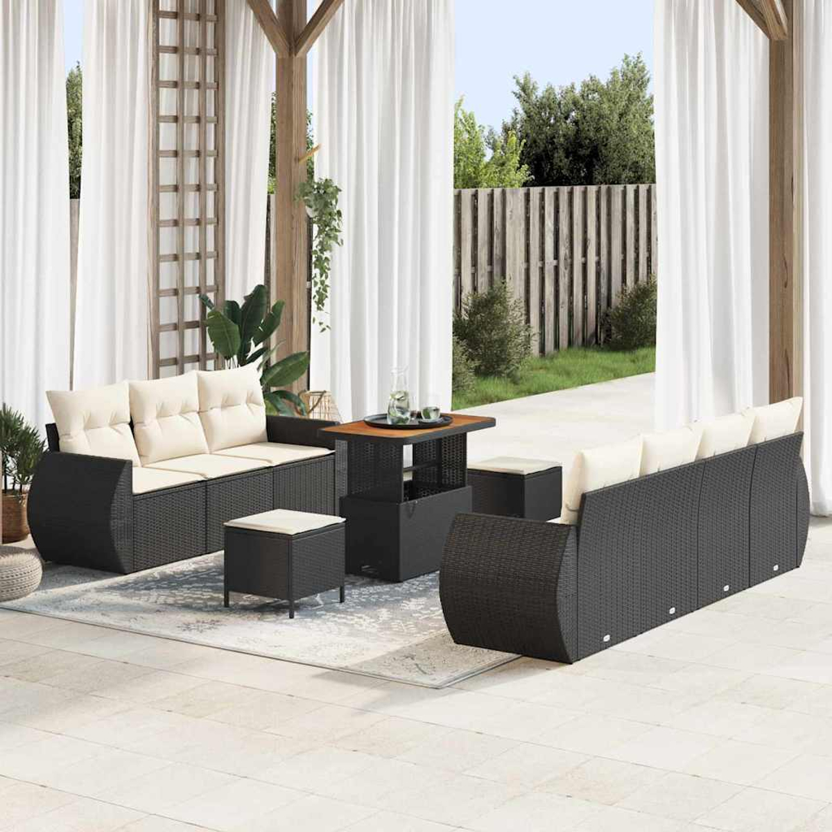 Juego de muebles de patio. Sofás y otomanas de mimbre negro con cojines crema. Mesa de madera. Cortinas blancas y jardín verde al fondo.