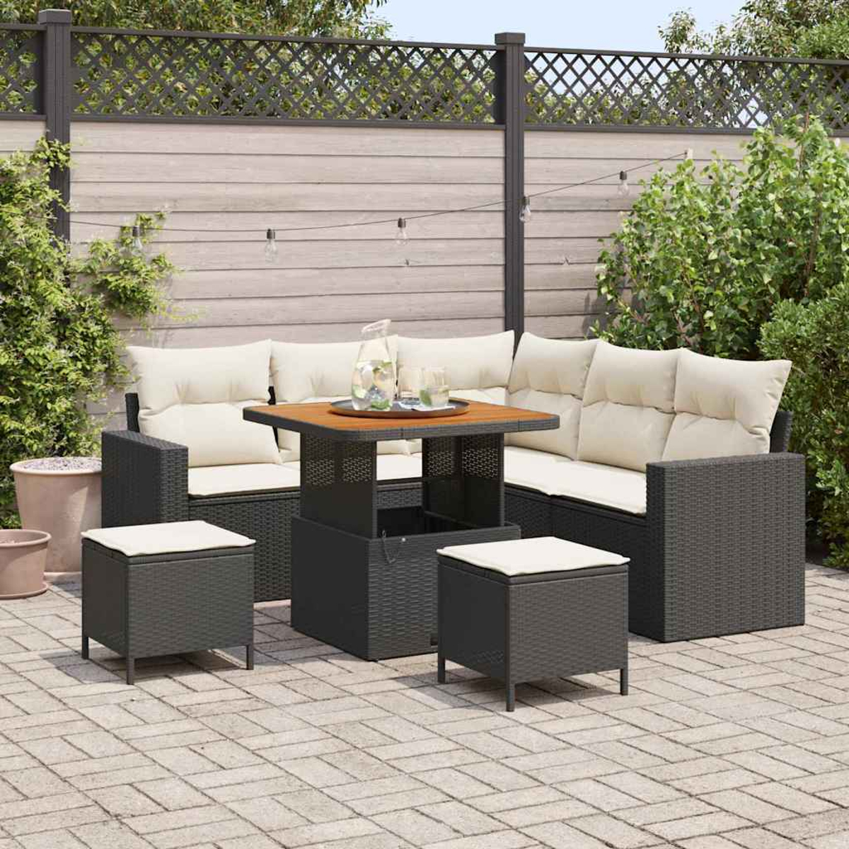Garten-Sitzgruppe mit schwarzem Korbgeflecht, cremefarbenen Kissen, Tisch und Hockern. Auf einer Ziegelterrasse mit Holzzaun im Hintergrund.