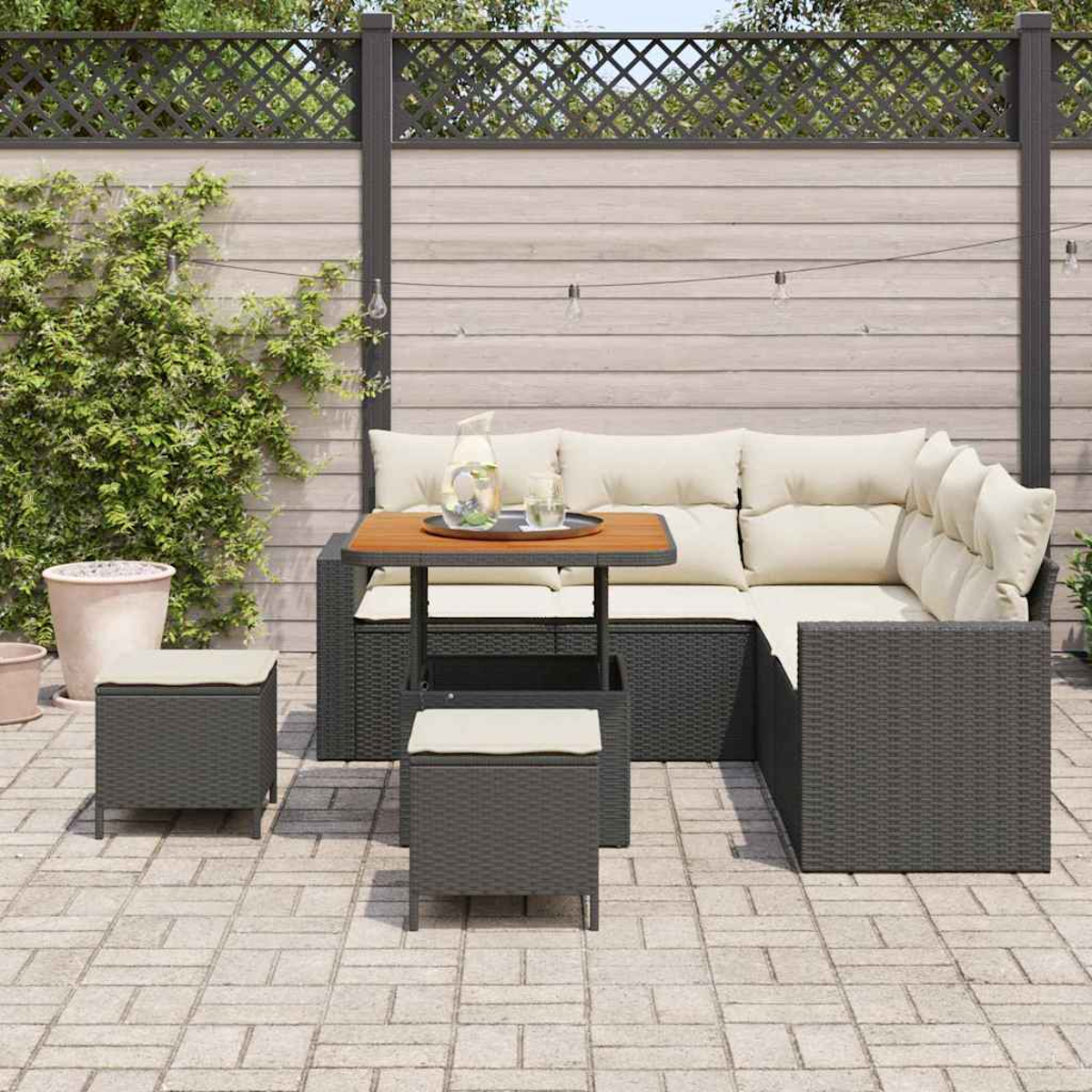 Gartenmöbel-Set. Dunkelgraues Korbsessel-Sofa mit cremefarbenen Kissen, kleinen Hockern und einem Holztisch. Dekorativer Zaun.