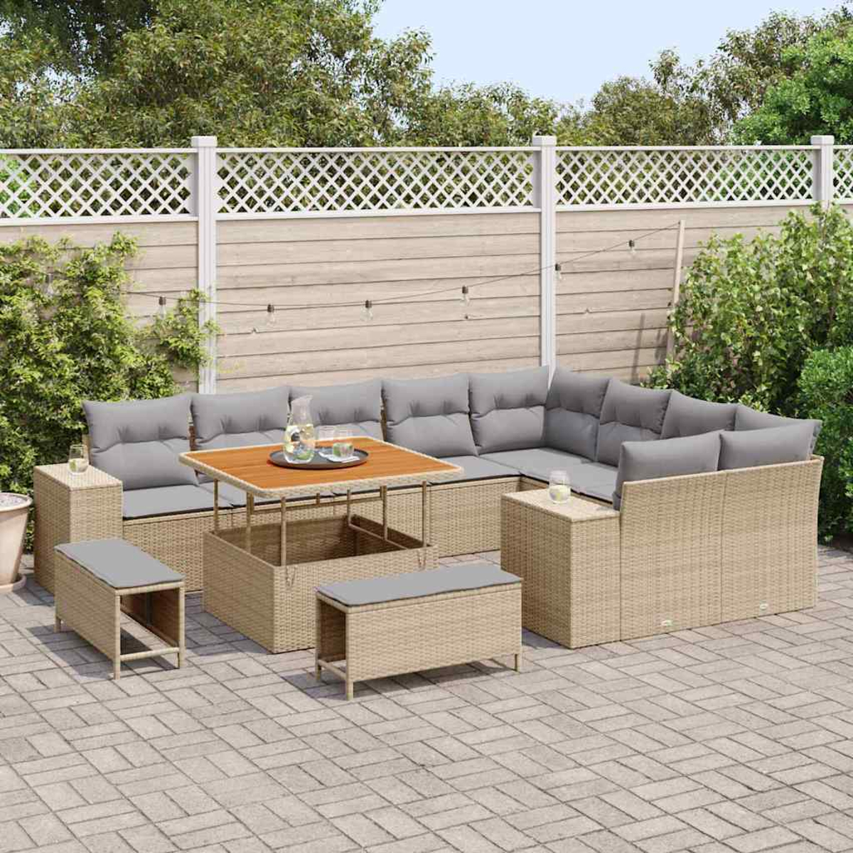 Set da patio esterno con mobili in vimini marrone chiaro, cuscini grigi e un tavolo in legno. Recinto bianco e vegetazione sullo sfondo.