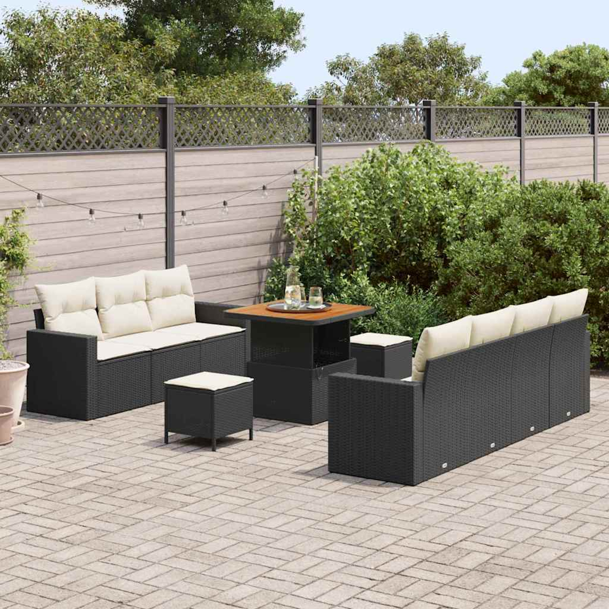 Terrassenmöbel-Set. Schwarze Korbmöbel mit cremefarbenen Kissen. Tisch mit Getränken. Steinpflaster mit Holzzaun und Grün.