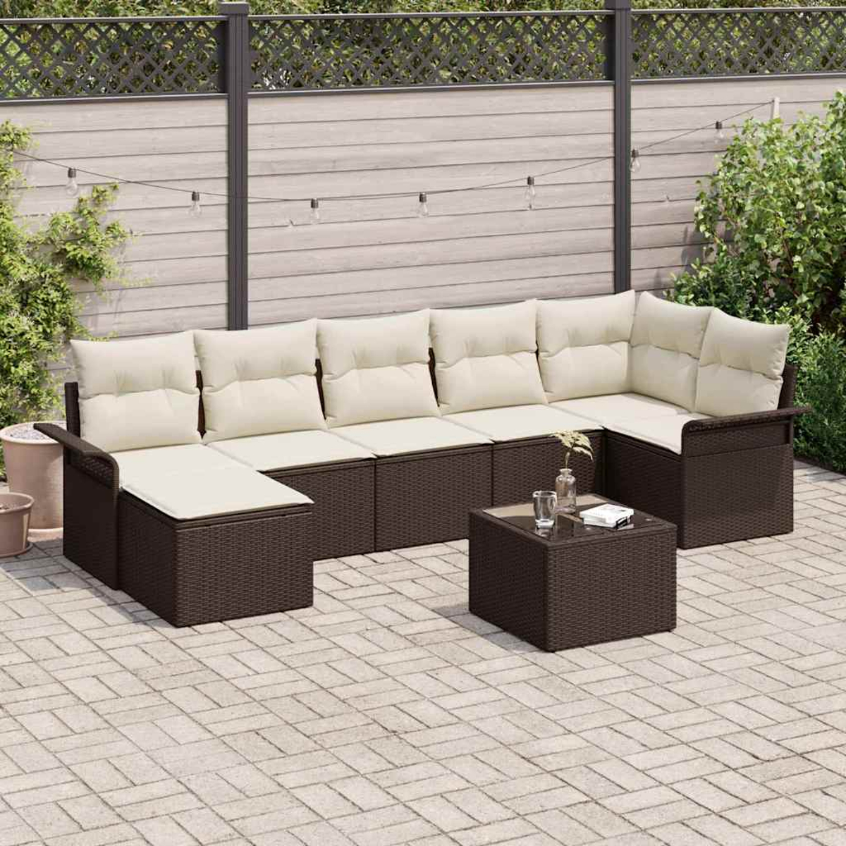 Outdoor-Ecksofa mit cremefarbenen Kissen und einem kleinen Tisch. Die Möbel sind dunkelbraun. Dahinter ein Holzzaun und Grün.