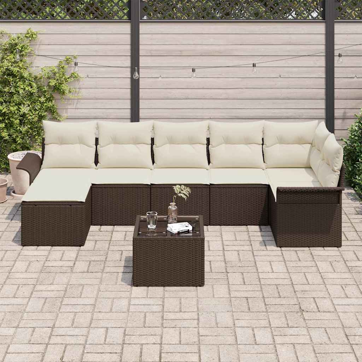 Garten-Sitzgruppe mit braunem Korbgewebe und cremefarbenen Kissen, kleiner Tisch davor. Auf einer Backstein-Terrasse mit Holzzaun.