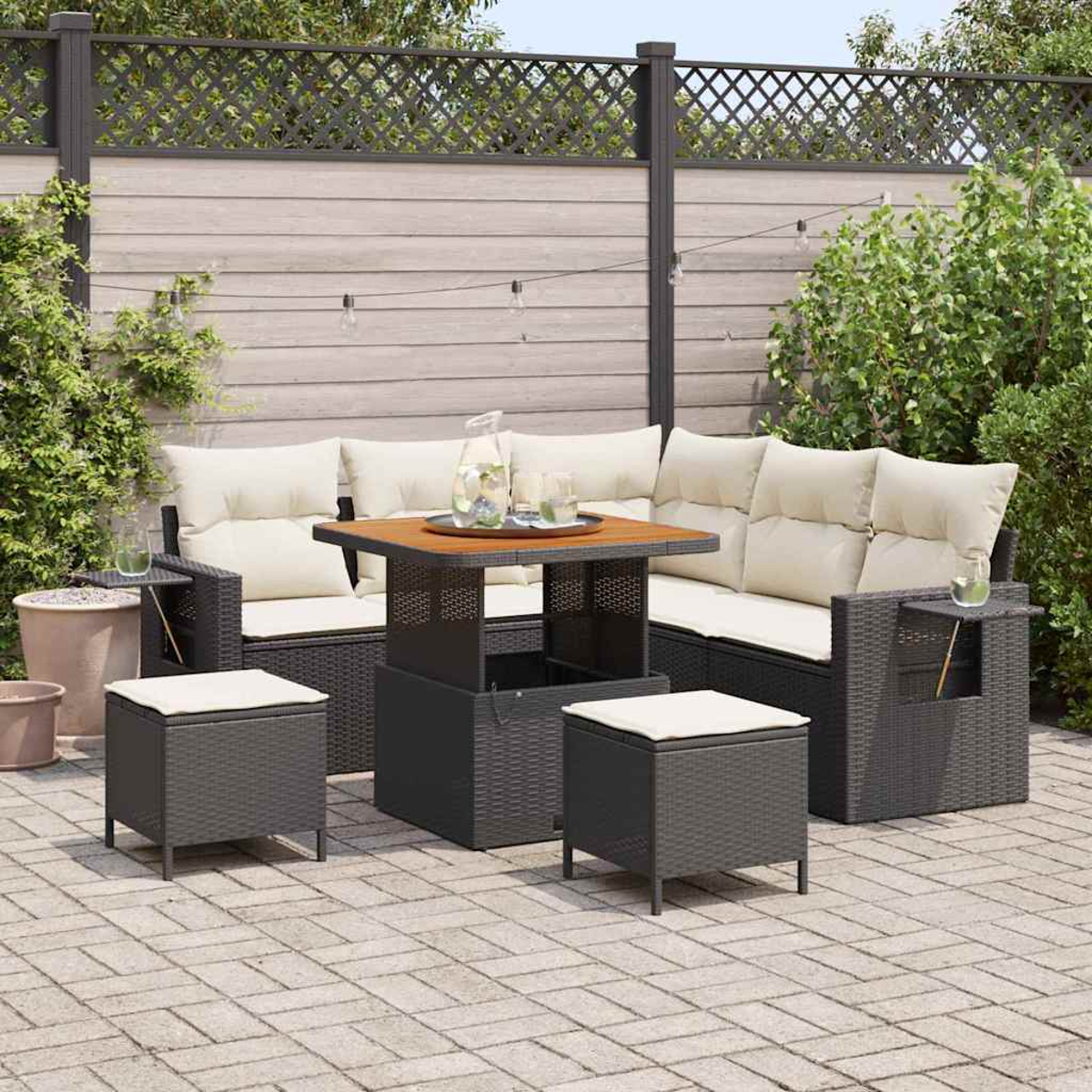 Set da patio esterno con mobili in vimini scuro, cuscini color crema e un tavolo quadrato. Include due pouf. Recinto di legno sullo sfondo.