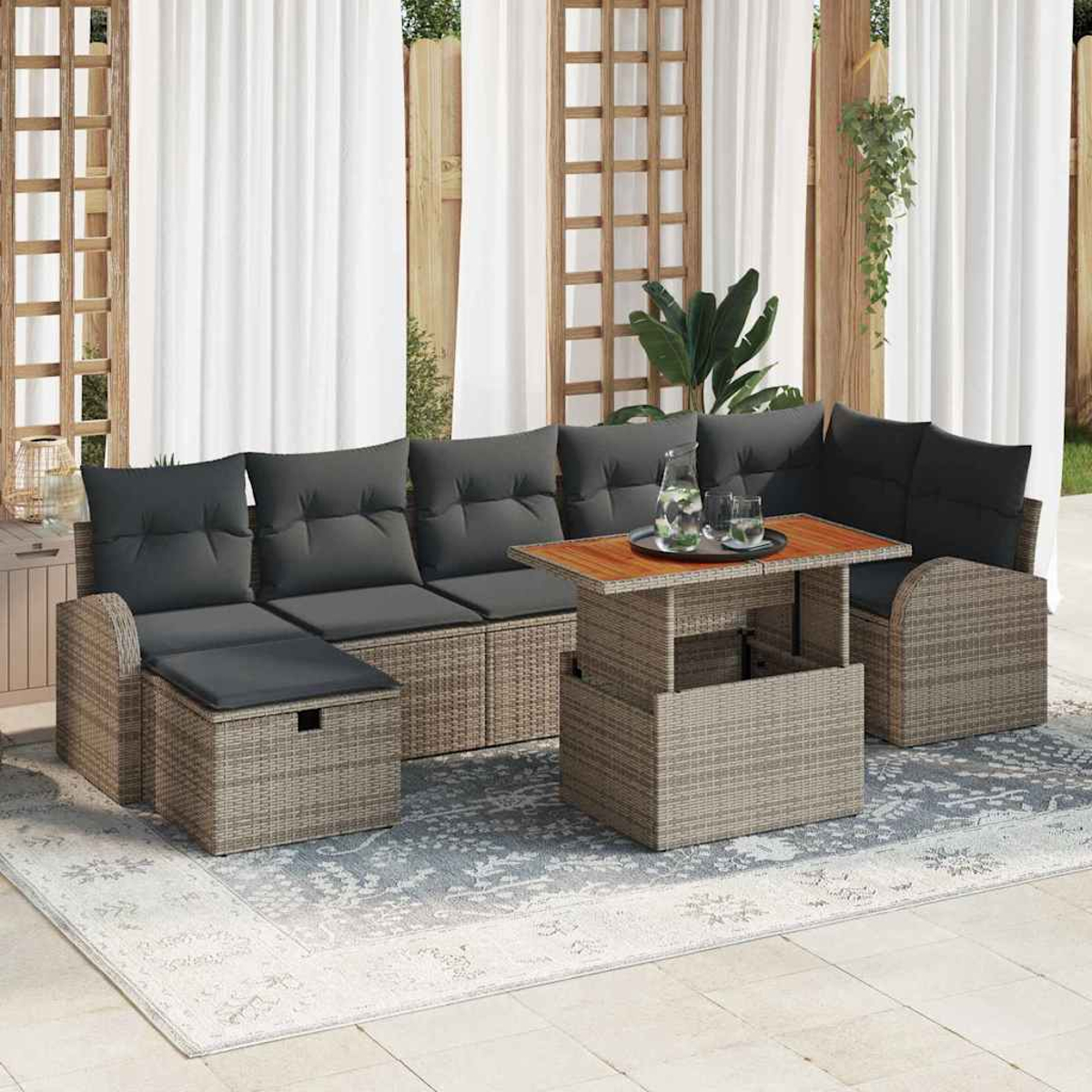 Gartenmöbel-Set aus Korbgeflecht mit grauen Kissen und einem Holztisch auf einem gemusterten Teppich, vor weißem Vorhang.