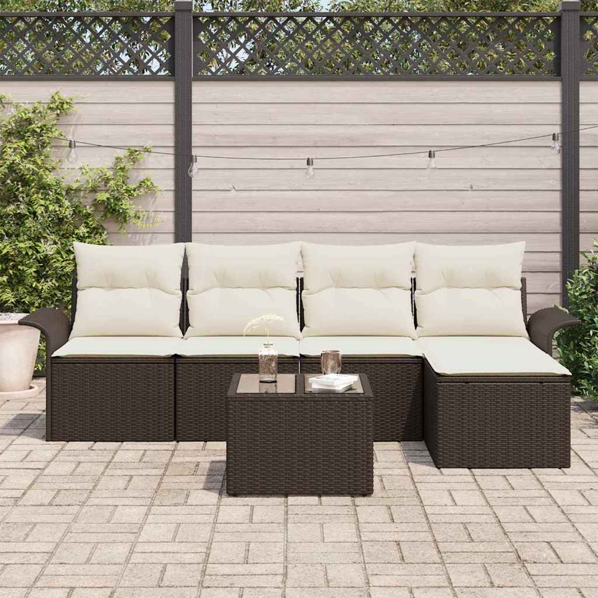 Gartenmöbel-Set mit braunem Korbgeflecht, cremefarbenen Kissen und einem kleinen Tisch mit Glasplatte auf einer Ziegelterrasse.